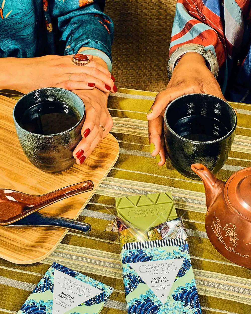 Compartés’ matcha green tea chocolate bar on a table, next to two tea cups being held by hands