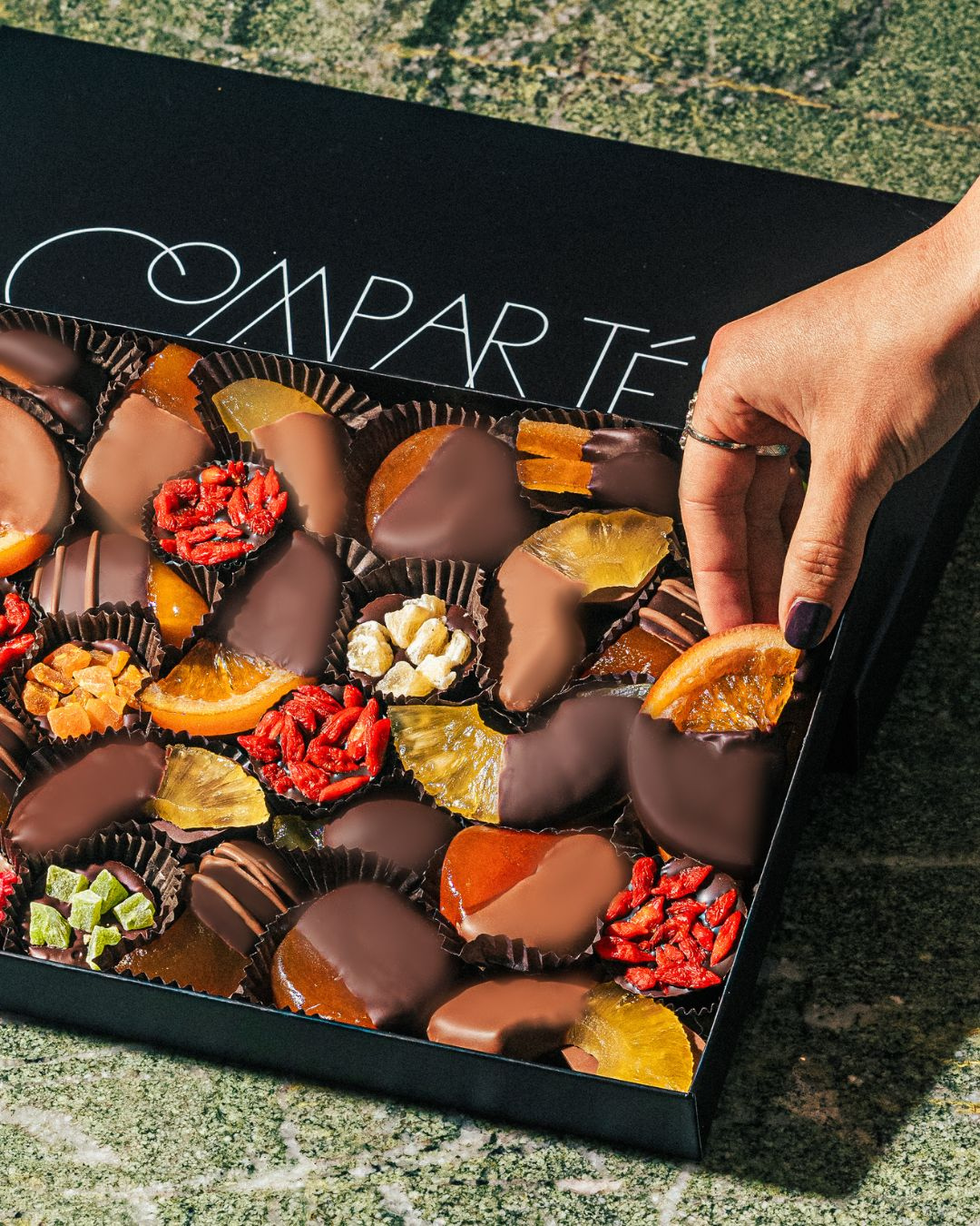 A hand grabbing milk chocolate, dark chocolate, and white chocolate-covered fruit in a large black gift box.