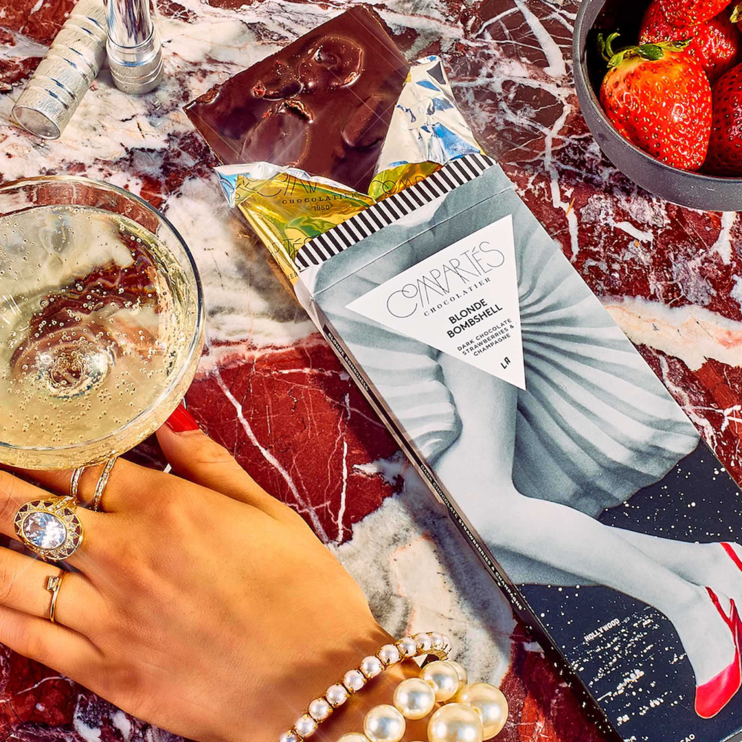 Compartés Blonde Bombshell chocolate bar styled on red marble with champagne, strawberries, and a jeweled hand.