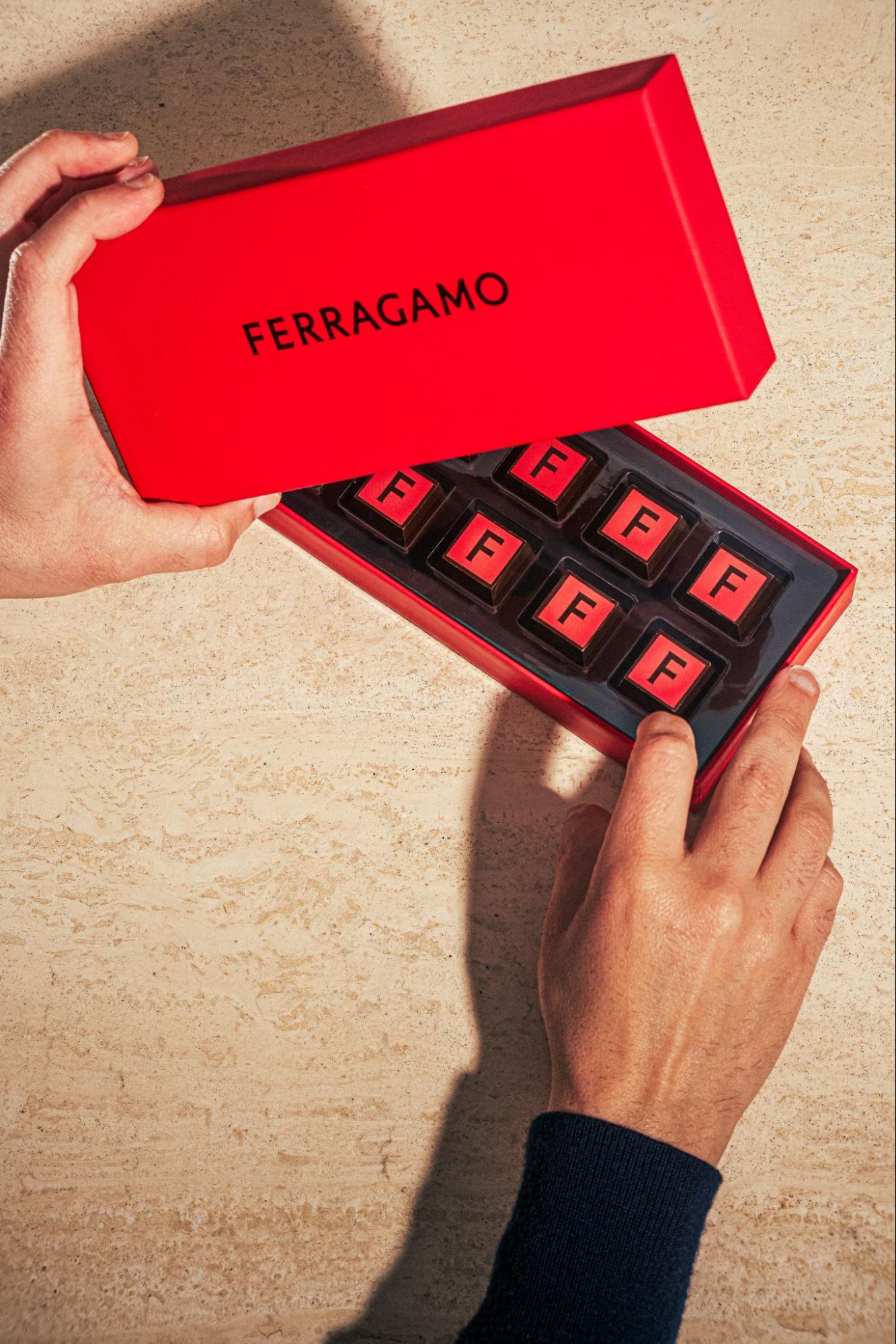 Hands opening a custom Compartés x Ferragamo branded chocolate gift box in signature red, revealing chocolates topped with the red "F" Ferragamo logo, a luxury corporate gift idea that goes beyond the ordinary.