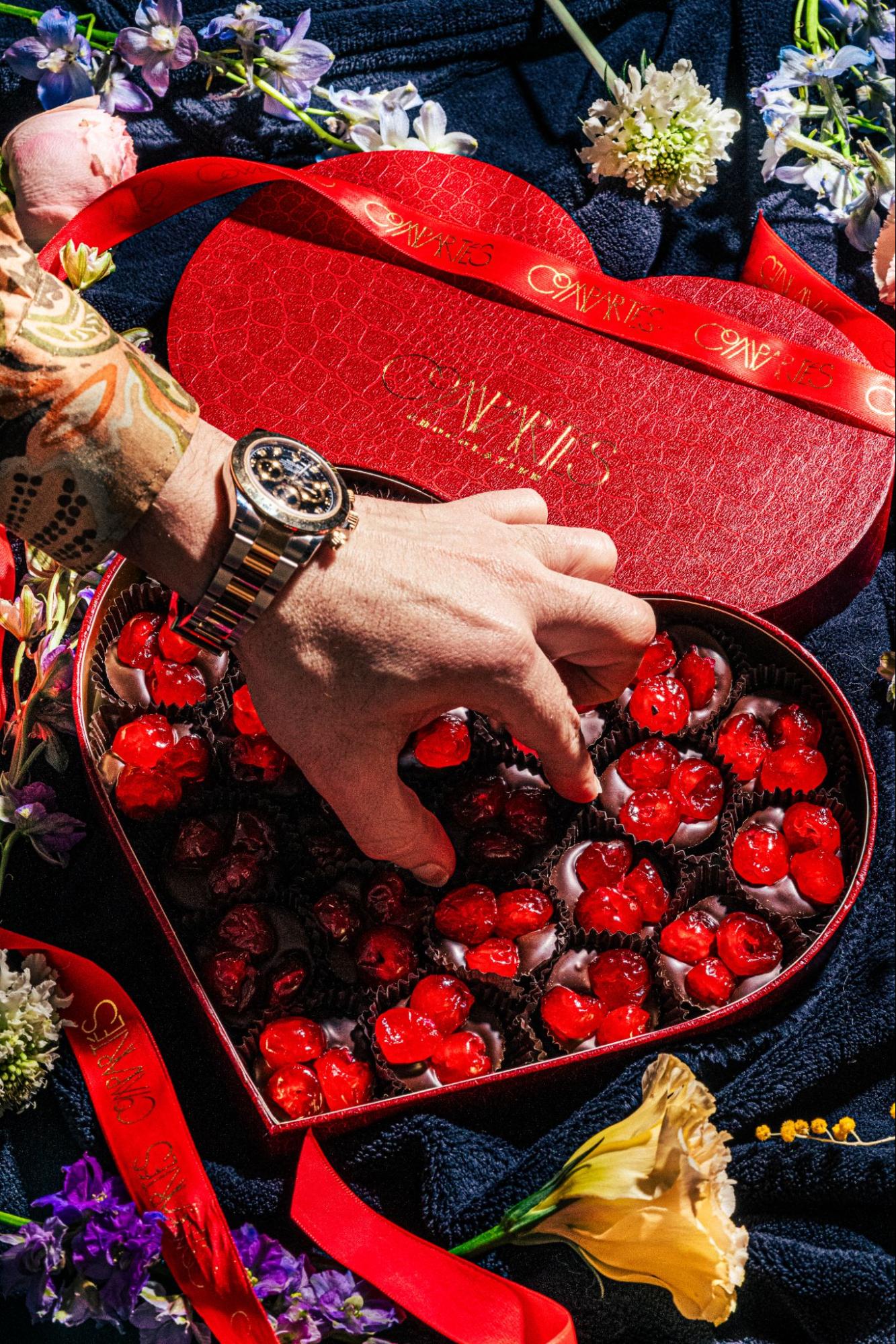 Hand reaching into heart shaped box of chocolate dipped cherries with flowers in background