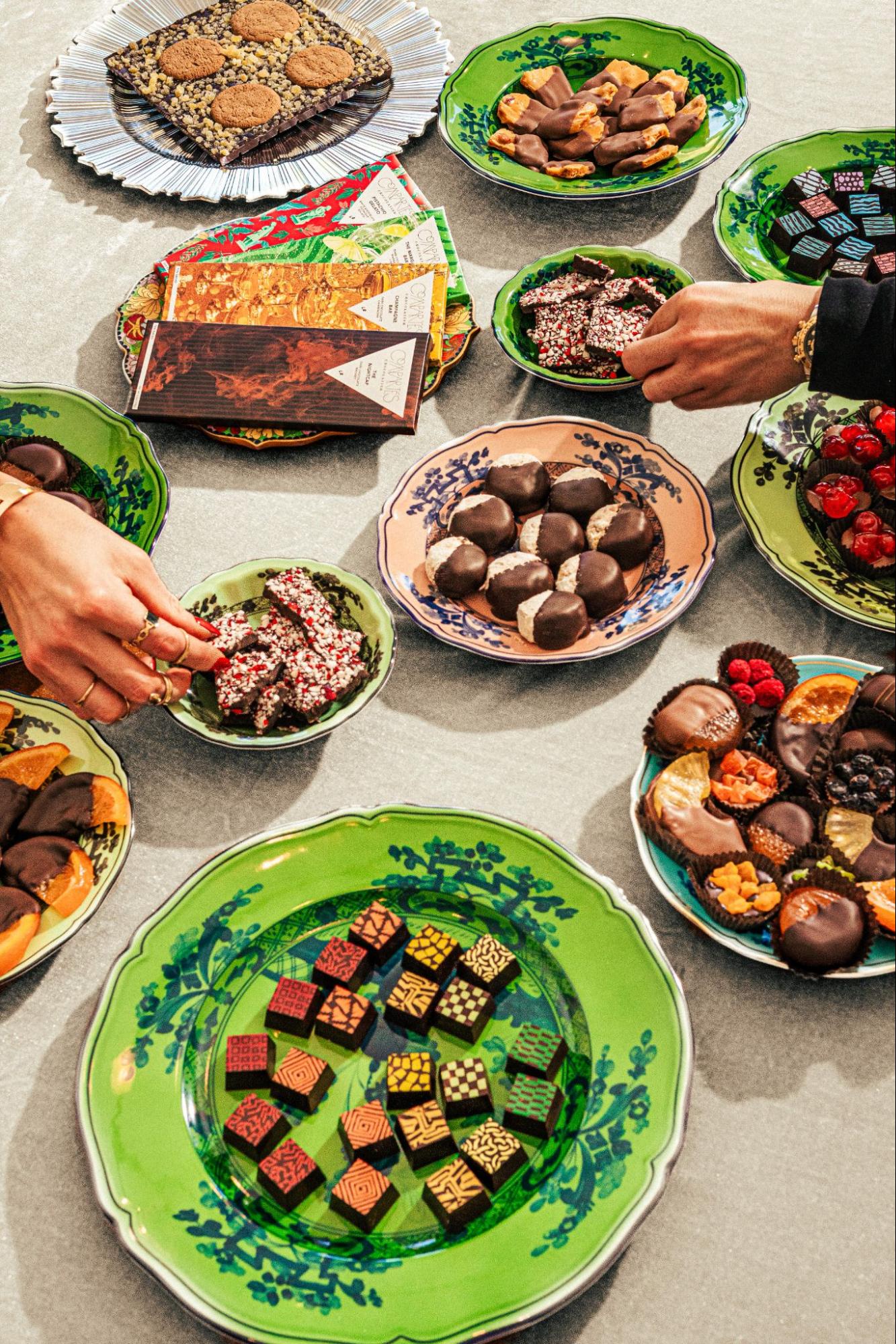 Spread of Compartés thank you chocolates on colorful patterned plates, featuring artistic printed bonbons, peppermint bark, chocolate-dipped fruit, toffee, and assorted holiday chocolate bars with hands reaching to share.