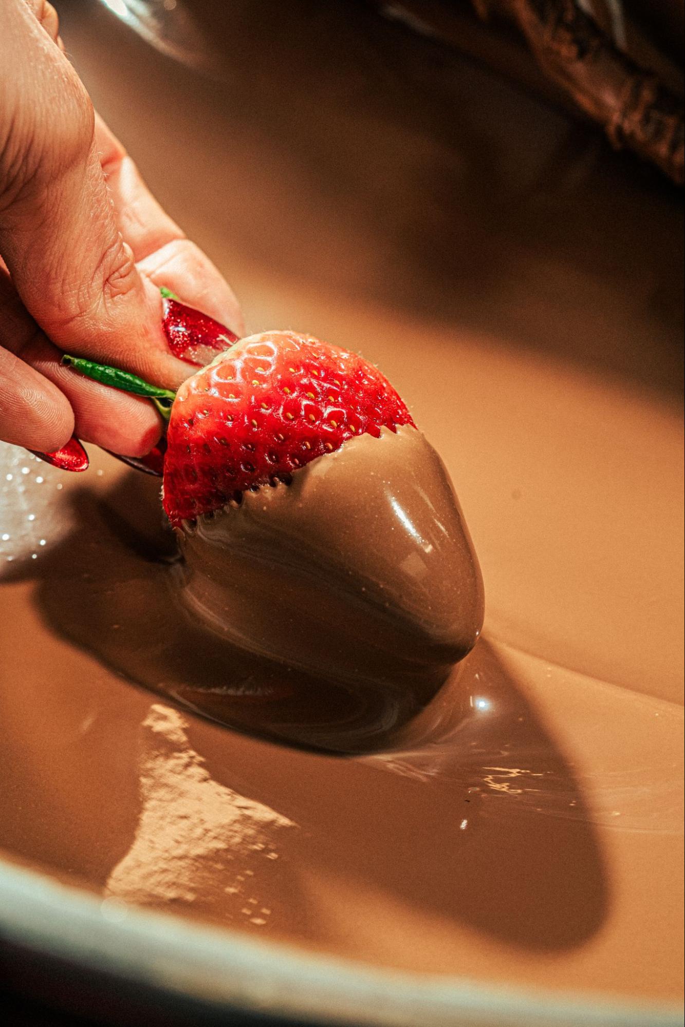 Hand dipping a fresh strawberry into a bowl of glossy tempered milk chocolate, showcasing the smooth, silky texture of perfectly tempered chocolate coating