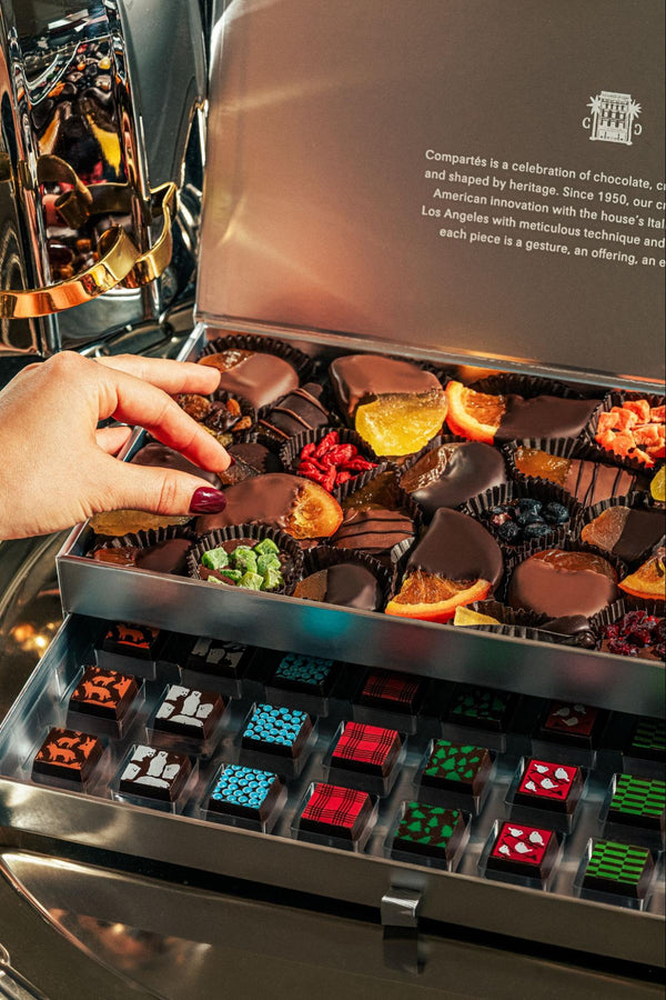 A hand selecting a chocolate-dipped citrus truffle from a luxury Compartés chocolate assortment displayed in a silver gift box with assorted artisan chocolates.