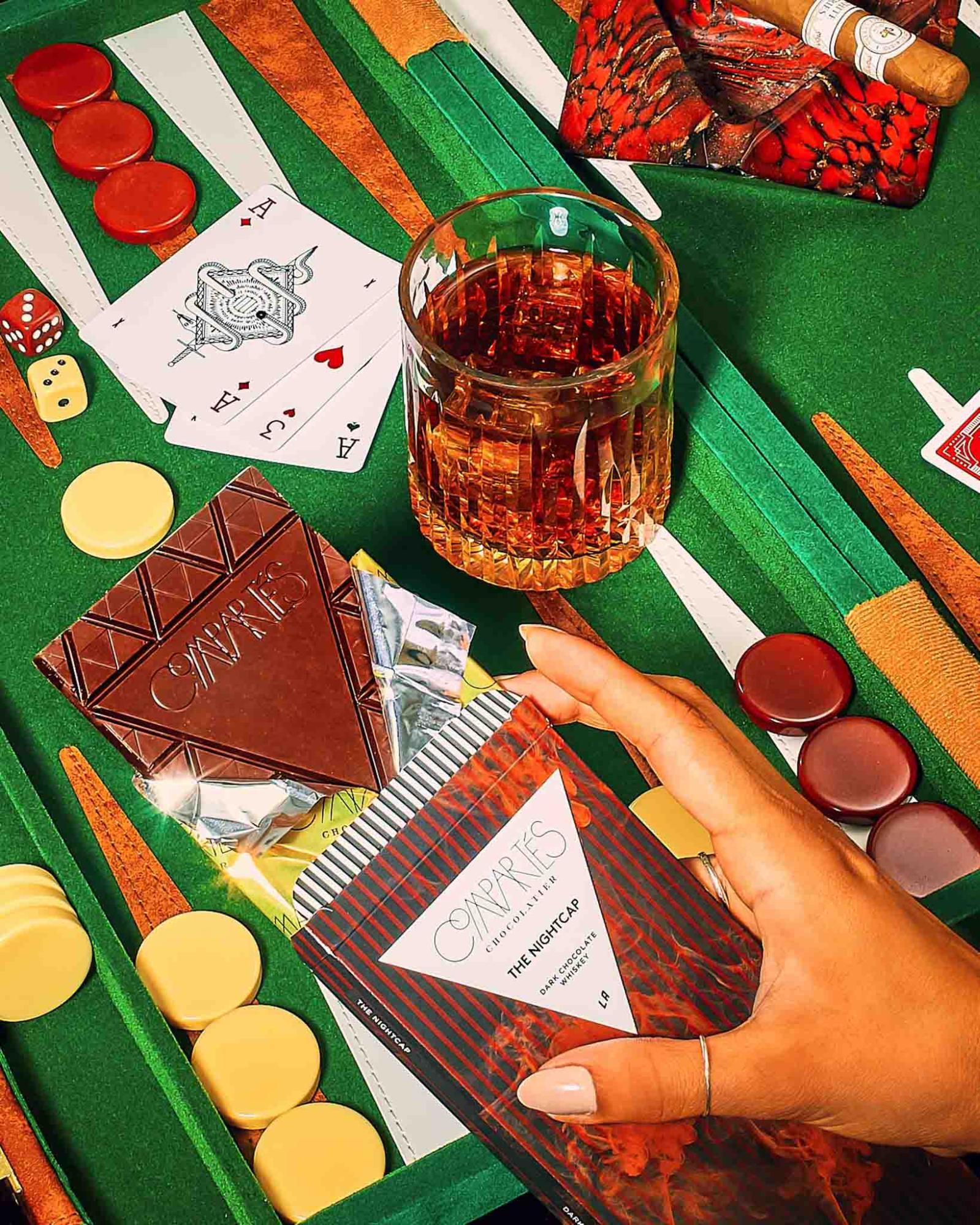 Luxury chocolate and whiskey pairing featuring gourmet dark chocolate bars, playing cards, poker chips, and a glass of whiskey on a casino-style game table