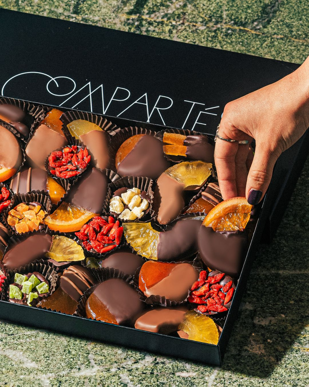 A hand grabbing milk chocolate, dark chocolate and white chocolate covered fruit in a large black gift box– Compartés