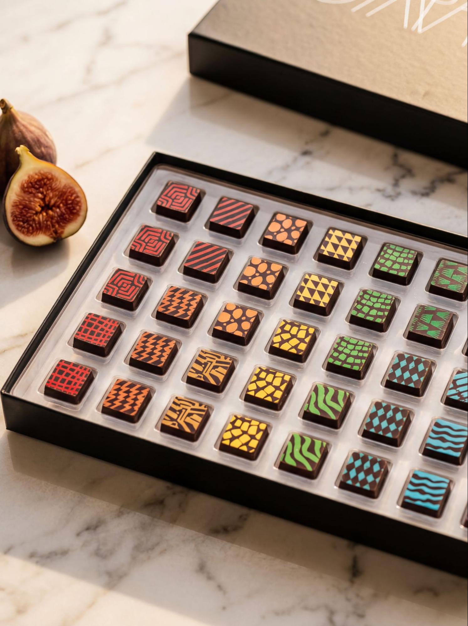 Compartés luxury gift box of colorful hand-painted artisan chocolate bonbons in rainbow-ordered patterns, styled with fresh figs on white marble as a sophisticated corporate gift idea.