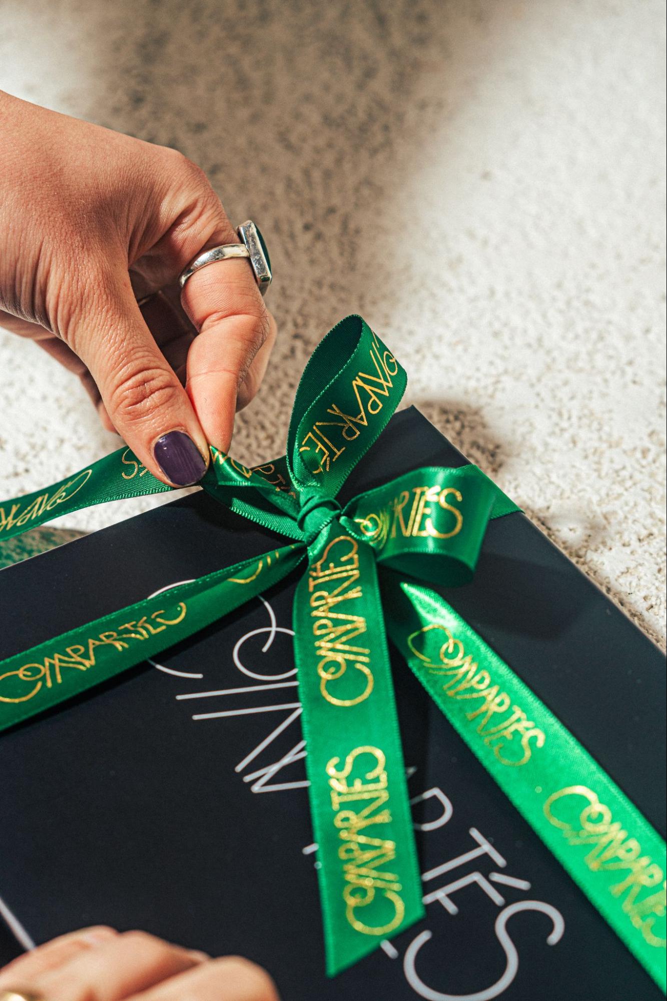 Hands tying a green Compartés ribbon with gold logo lettering into a bow on a black gift box, illustrating branded corporate gifting and premium gift presentation for modern brands.