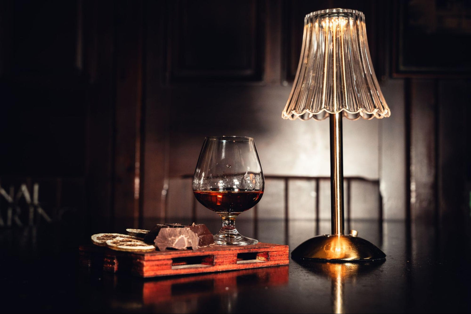 A cordless crystal table lamp with a pleated clear shade and a metallic gold base is next to a glass of whiskey, dried citrus fruits, and dark chocolate.