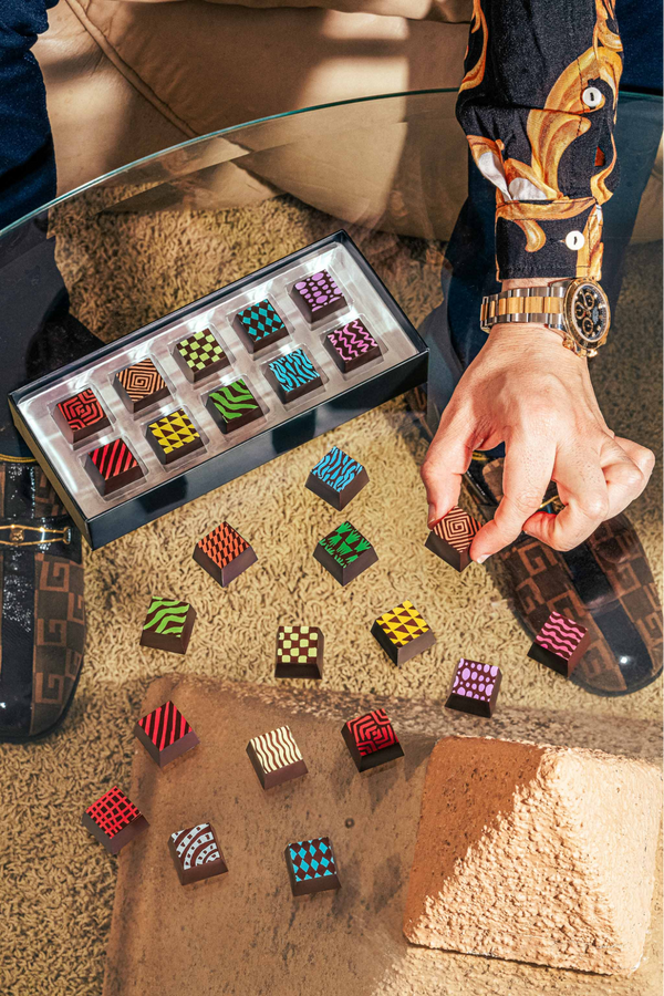 A man wearing a luxury watch and patterned shirt reaches for a square chocolate from an arrangement of treats with neon geometric patterns.