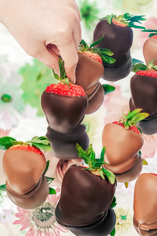 A hand reaches for a dark chocolate-dipped strawberry from a floating arrangement against a floral background.