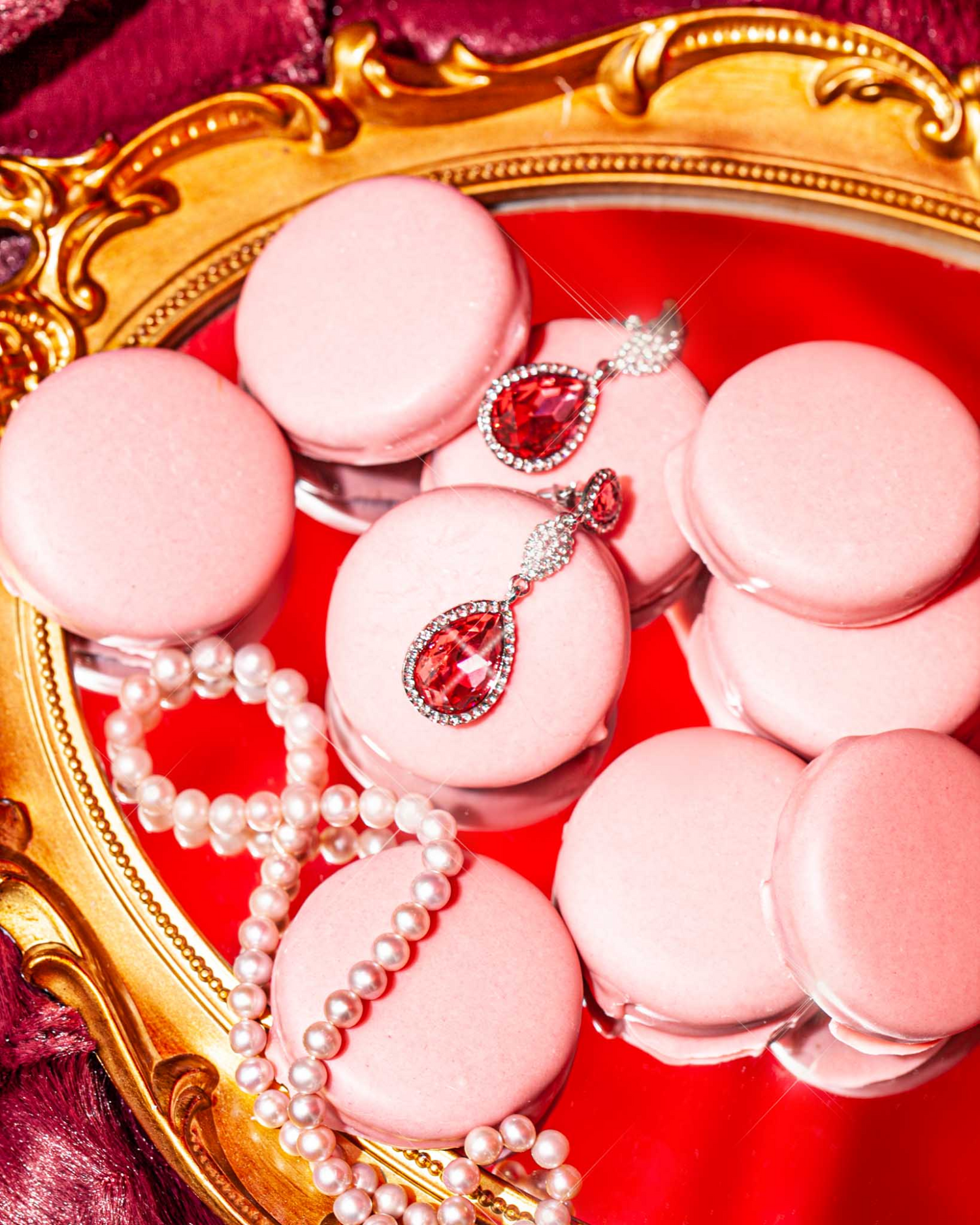 A pair of pear-shaped red gemstone earrings and a pearl necklace resting on pink chocolate-covered Oreos atop a gold-framed mirror.