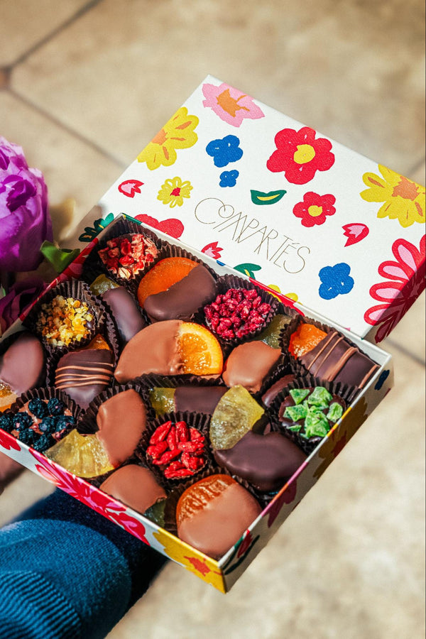 A woman in blue sweater holding a box of Compartés truffles & assorted chocolate fruit in a colorful floral box