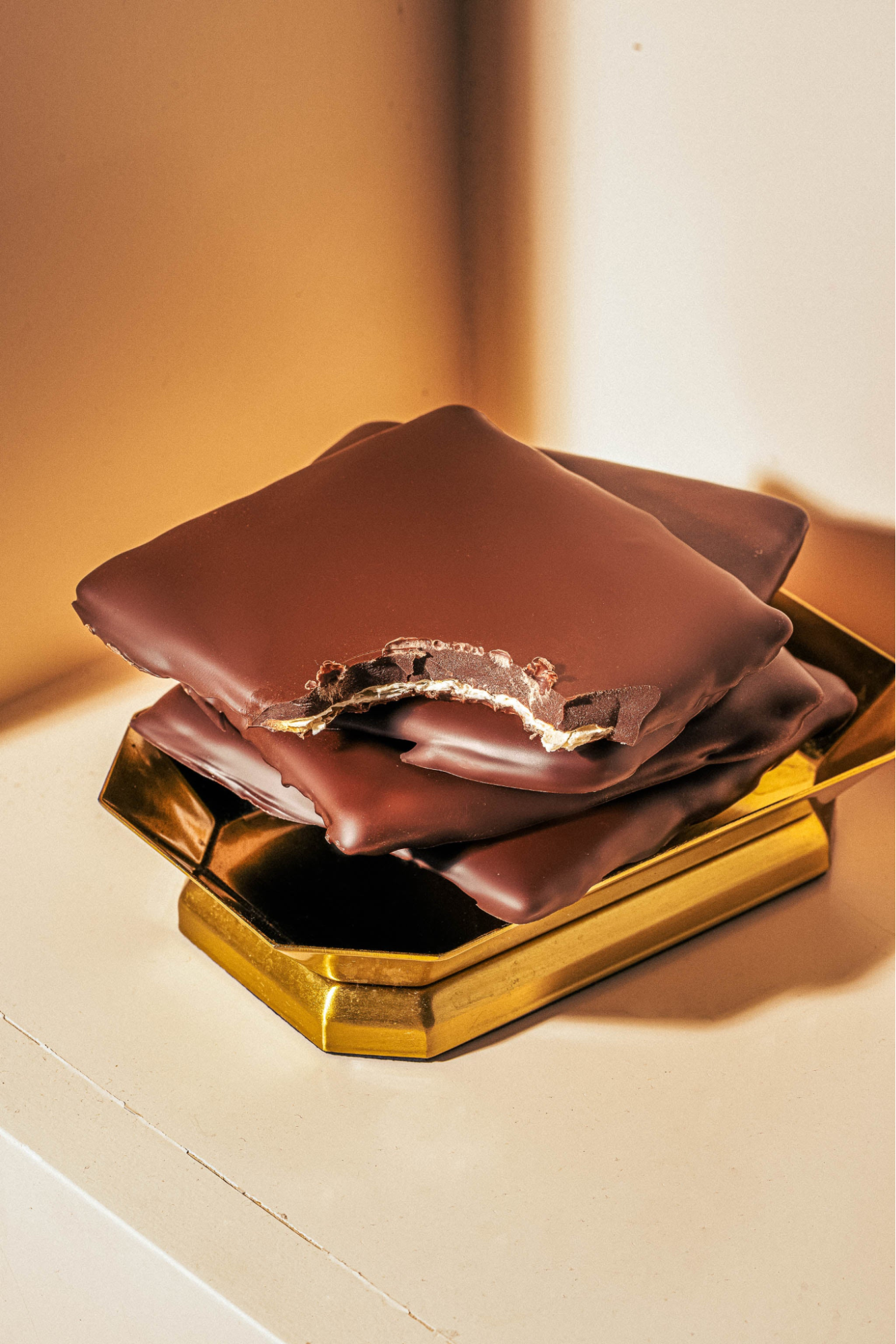 A stack of thin, dark chocolate-covered matzo treats on a gold tray, with the top piece showing a bite taken out of it.