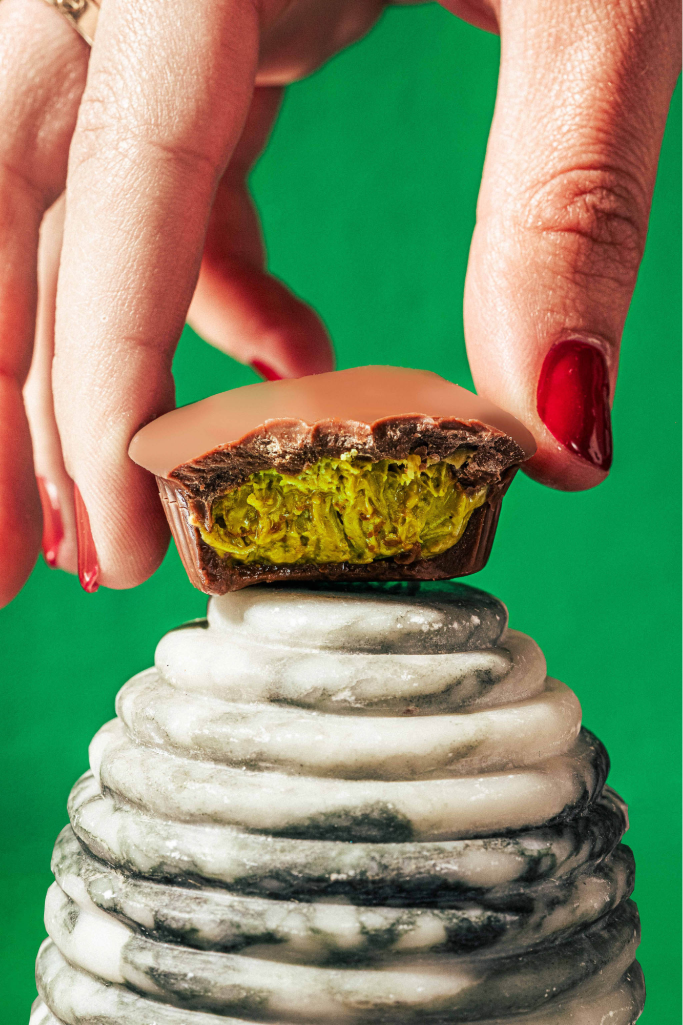 A close-up shot of a hand with red nail polish holding a bitten chocolate cup with a vibrant green, textured pistachio and knafeh filling, resting on a circular marble pedestal.