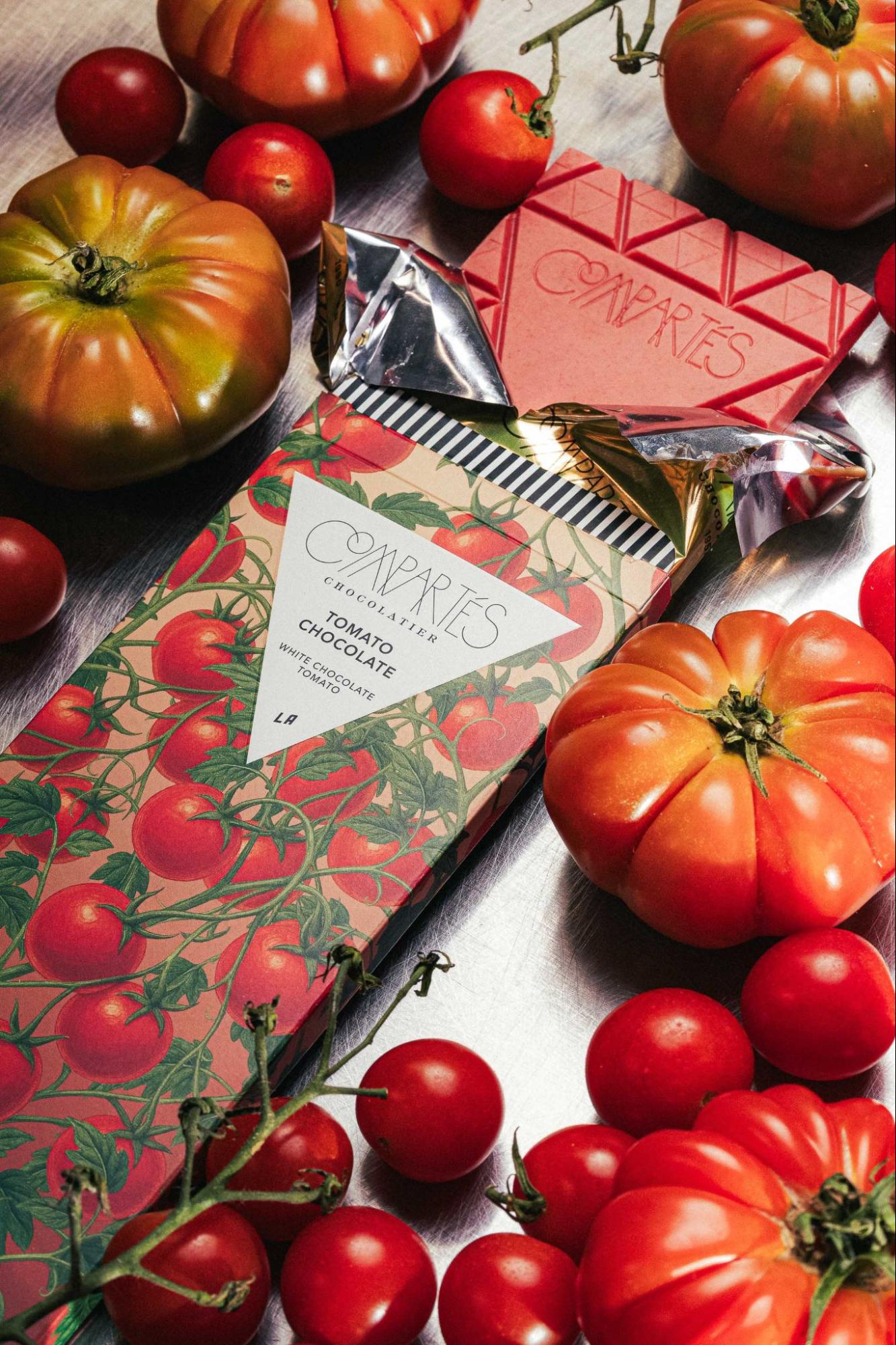 Compartés Tomato Chocolate bar, a weird chocolate flavor made with white chocolate and tomato, surrounded by fresh heirloom and cherry tomatoes on a silver tray with the pink-hued unwrapped bar visible.