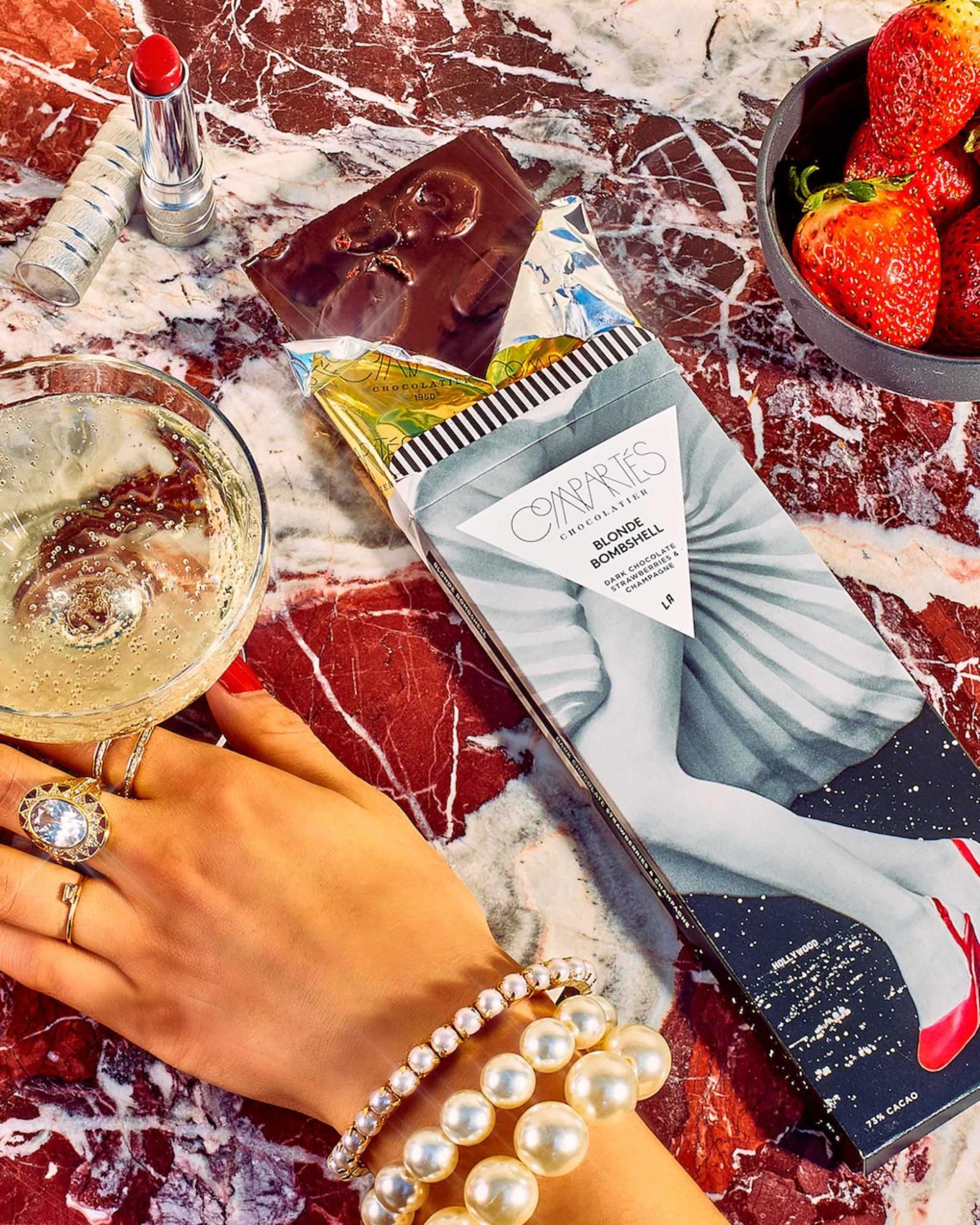 Compartés Blonde Bombshell dark chocolate bar with strawberries and champagne, styled with a champagne coupe, fresh strawberries, red lipstick, and pearl jewelry on red marble as an elegant chocolate pairing.