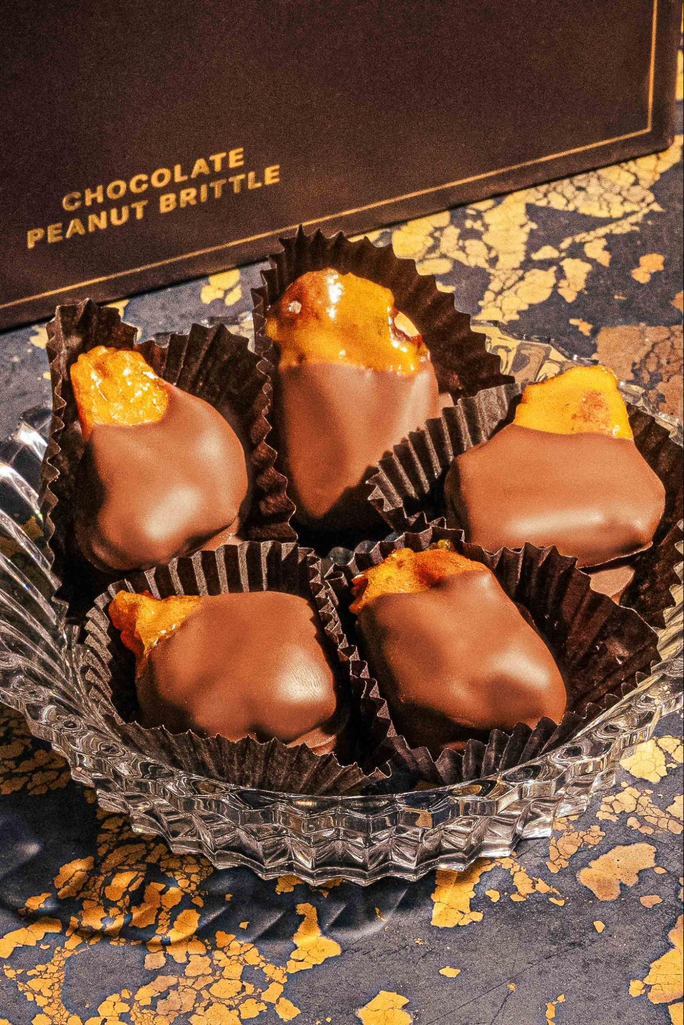 Compartés Chocolate Peanut Brittle pieces dipped in milk chocolate, displayed in dark cupcake liners on a crystal dish beside the gold-lettered gift box, a lavish chocolate dessert idea on a black and gold surface.