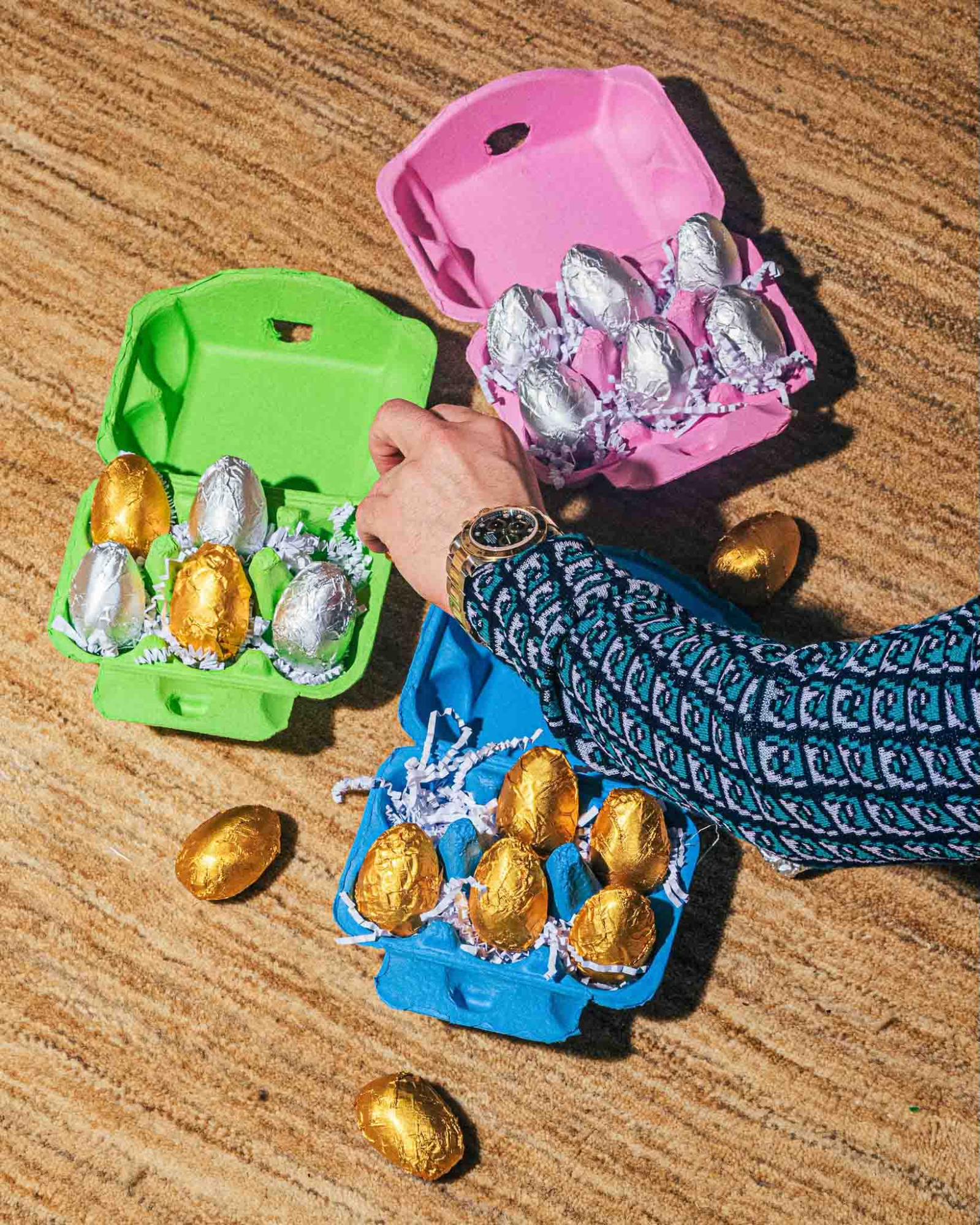 A reaches into a lime green egg carton filled with gold and silver foil-wrapped eggs, which sits next to matching pink and blue cartons on a textured tan surface.