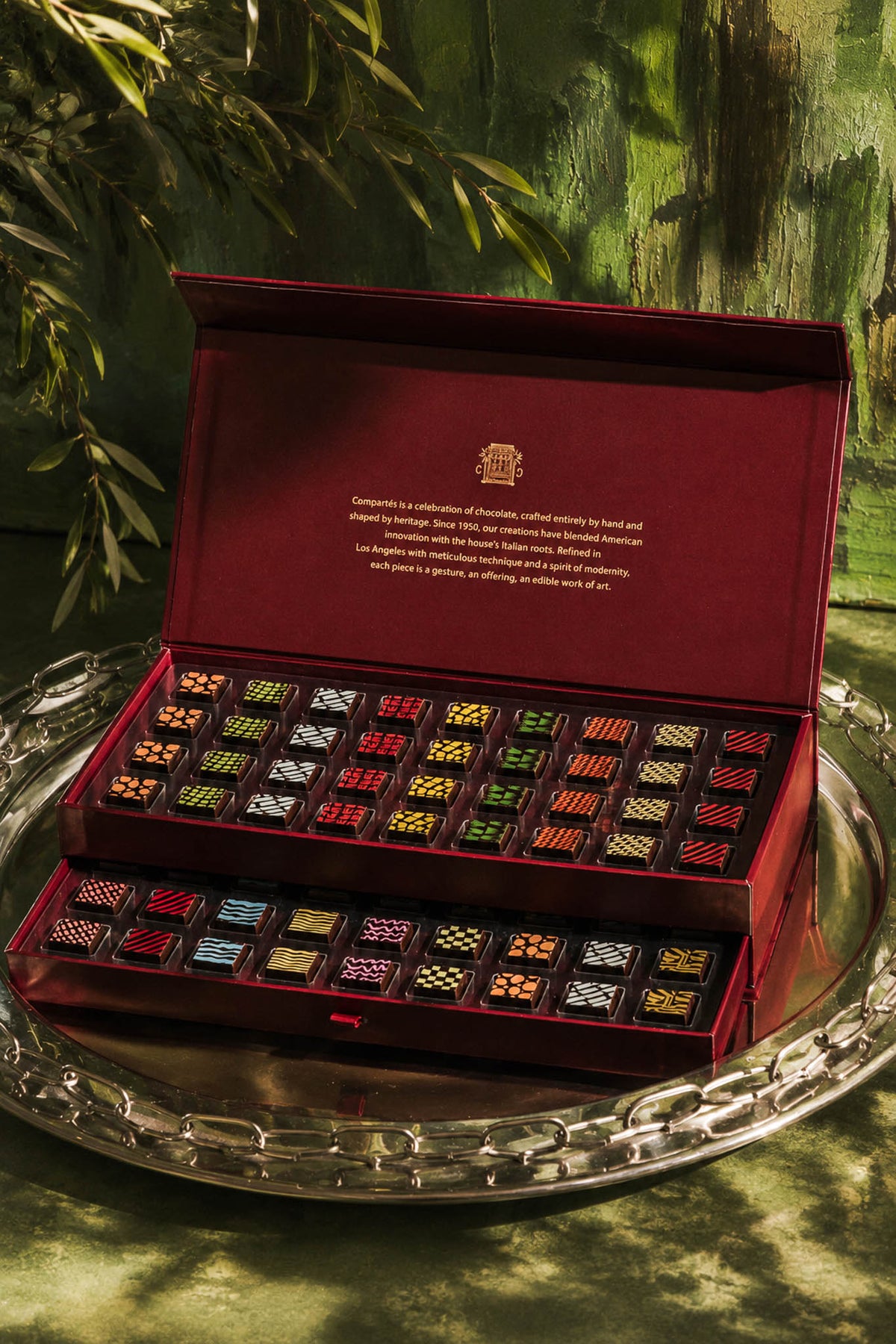 An open dark red luxury chocolate gift box displayed on a silver tray, with rows of assorted chocolates and chocolate truffles visible inside. The background is a painted green scene with foliage.