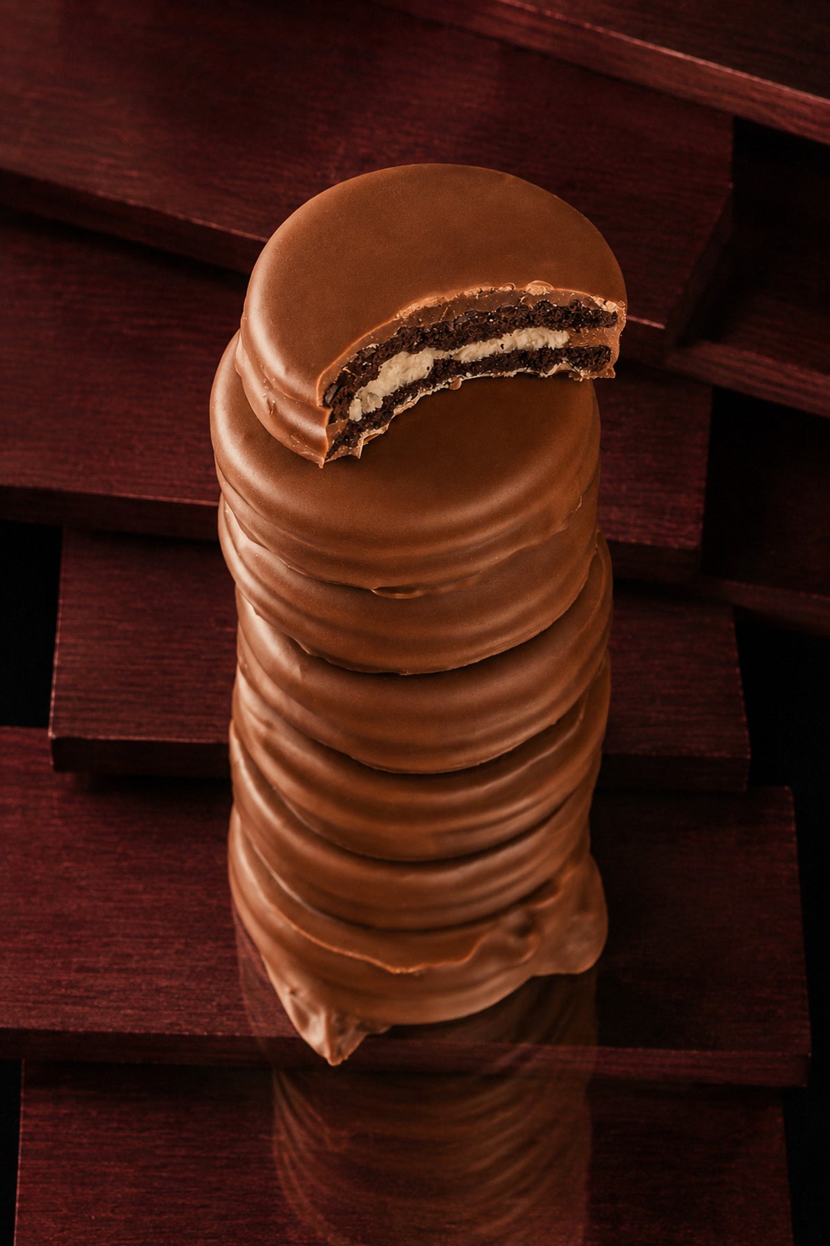 A stack of chocolate-covered Oreos cookies with a bite taken out at the top, showing a dark cookie and white cream filling, set against a dark wooden background.