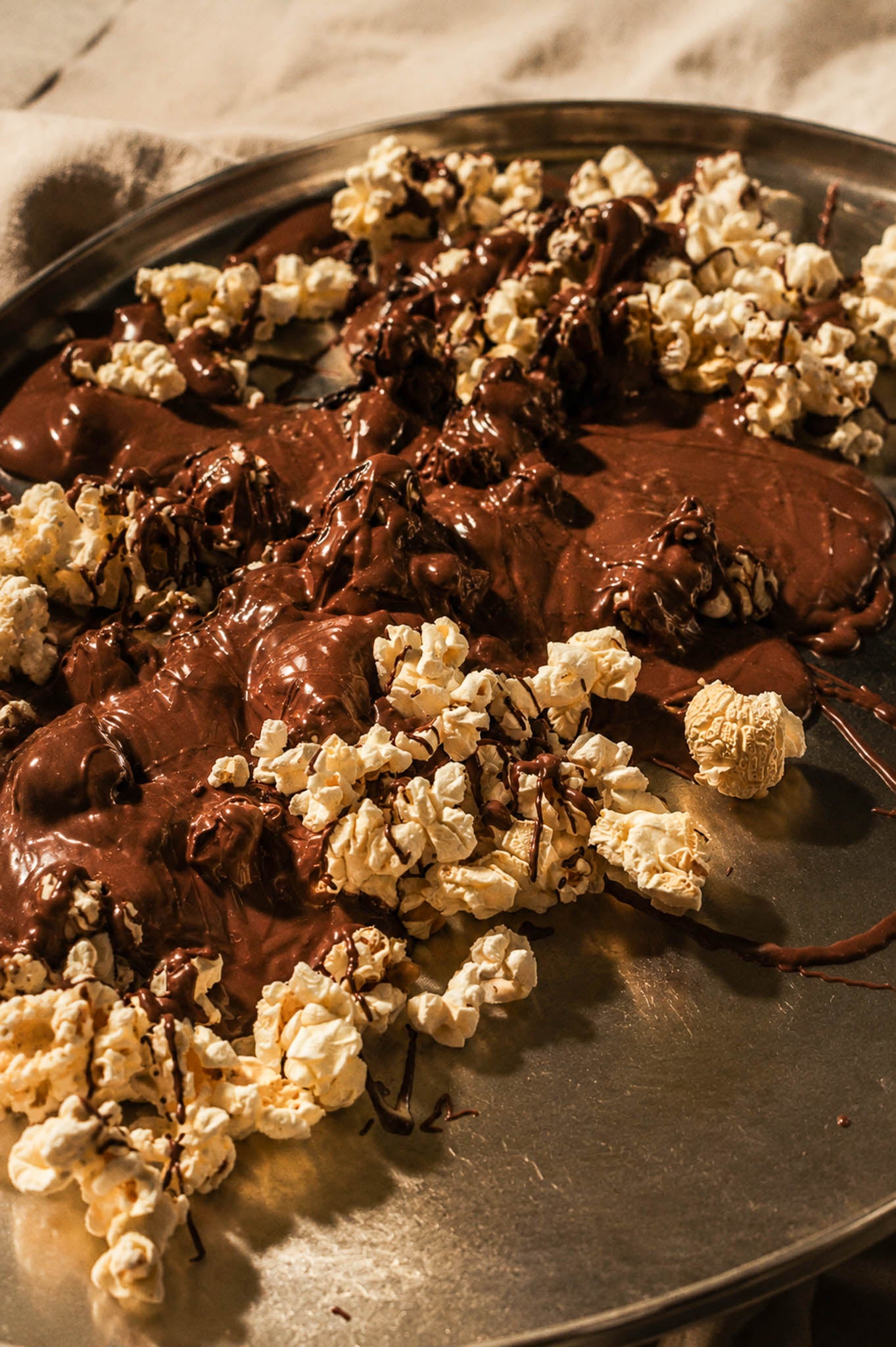 A close-up of gourmet chocolate popcorn mixed with melted dark chocolate on a round metal tray, photographed in warm, moody lighting.