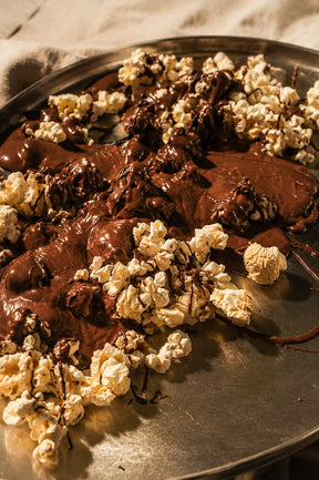 A close-up of gourmet chocolate popcorn mixed with melted dark chocolate on a round metal tray, photographed in warm, moody lighting.
