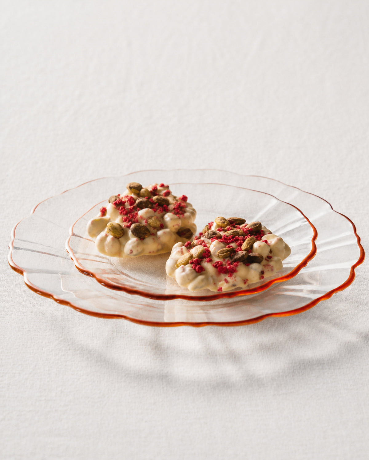 Chocolate with raspberry and green pistachios, served on a clear scalloped glass plate with a warm orange-brown rim on a white textured surface.
