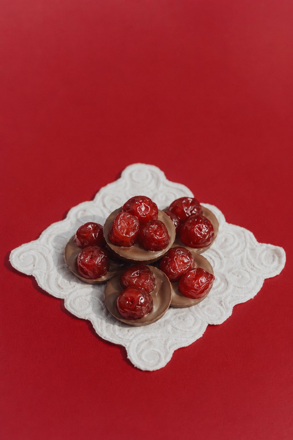 Gourmet luxury chocolate cherries with glossy red cherries, arranged on a white scalloped doily over a solid red background.