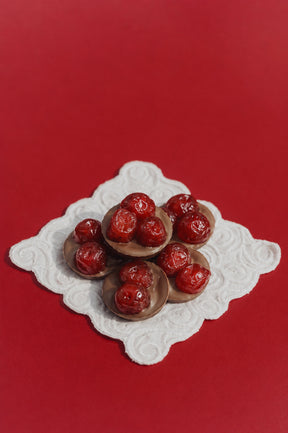 Gourmet luxury chocolate cherries with glossy red cherries, arranged on a white scalloped doily over a solid red background.