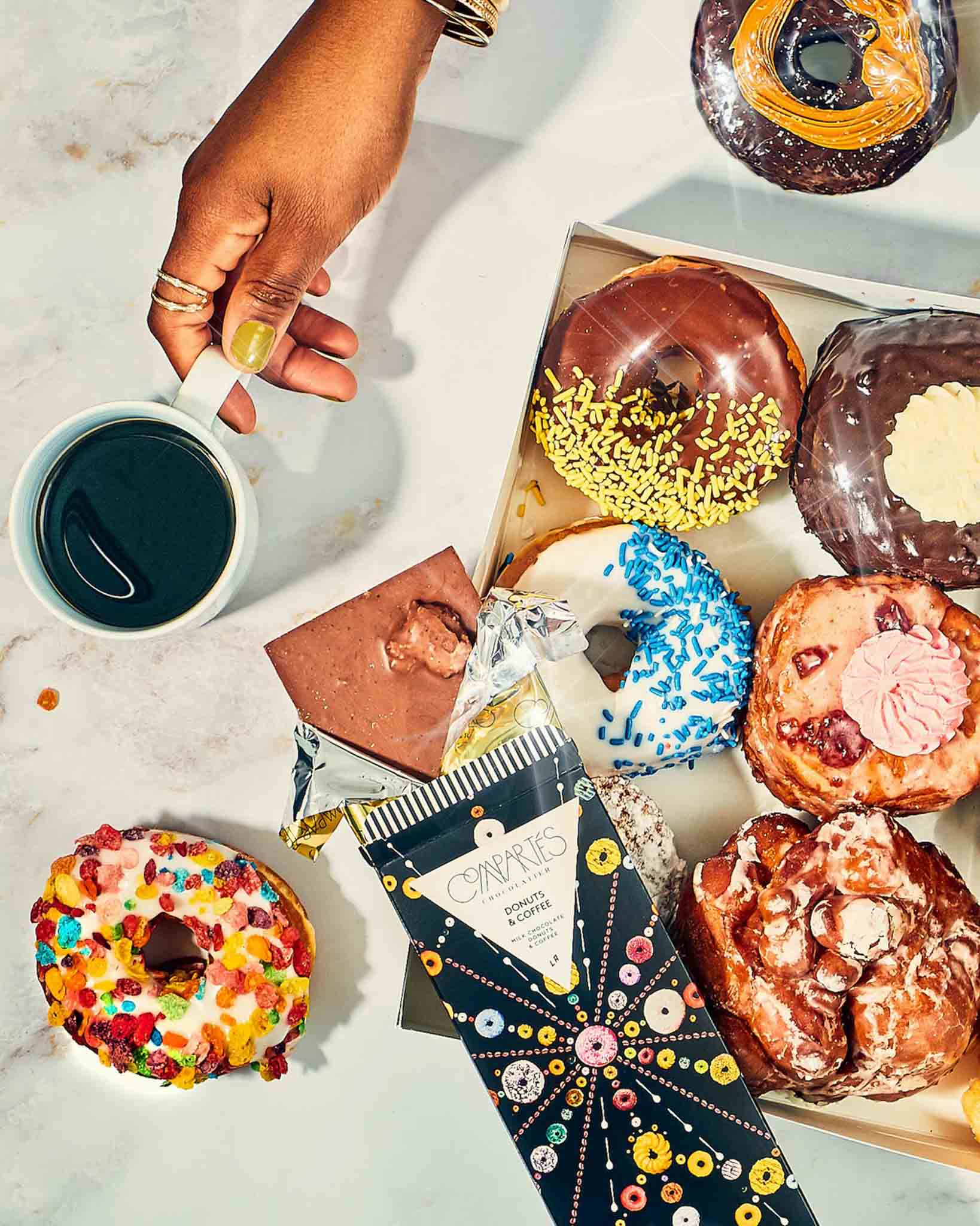 Overhead view of a box of donuts, a hand holding a cup of coffee, and the Coffee and Donuts Gourmet Chocolate Bar by Compartes